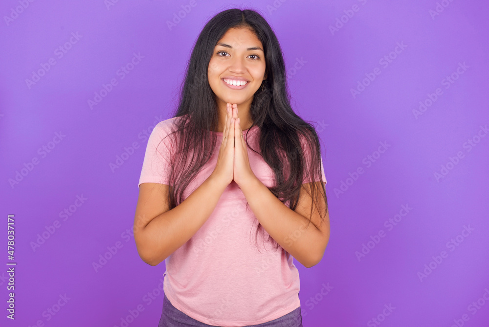 Hispanic brunette girl wearing pink t-shirt over purple background praying with hands together asking for forgiveness smiling confident. Hispanic brunette girl wearing pink t-shirt over purple background praying with hands together asking for forgiveness smiling confident.