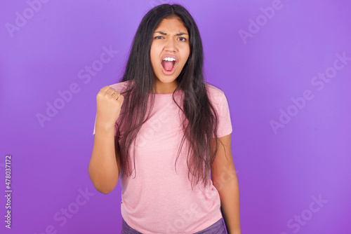Hispanic brunette girl wearing pink t-shirt over purple background angry and mad raising fist frustrated and furious while shouting with anger. Rage and aggressive concept.