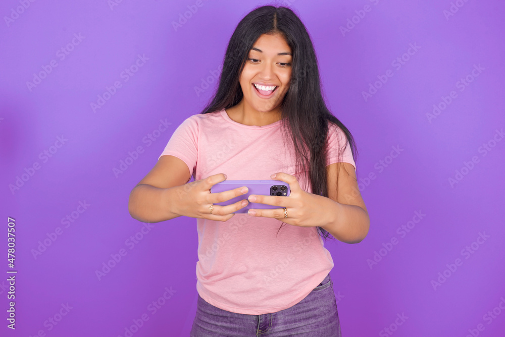 Portrait of an excited Hispanic brunette girl wearing pink t-shirt over purple background playing games on mobile phone. Portrait of an excited Hispanic brunette girl wearing pink t-shirt over purple background playing games on mobile phone.