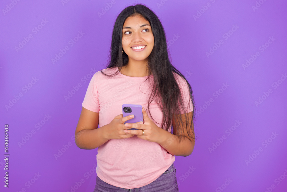 Hispanic brunette girl wearing pink t-shirt over purple background hold telephone hands read good youth news look empty space advert Hispanic brunette girl wearing pink t-shirt over purple background hold telephone hands read good youth news look empty space advert