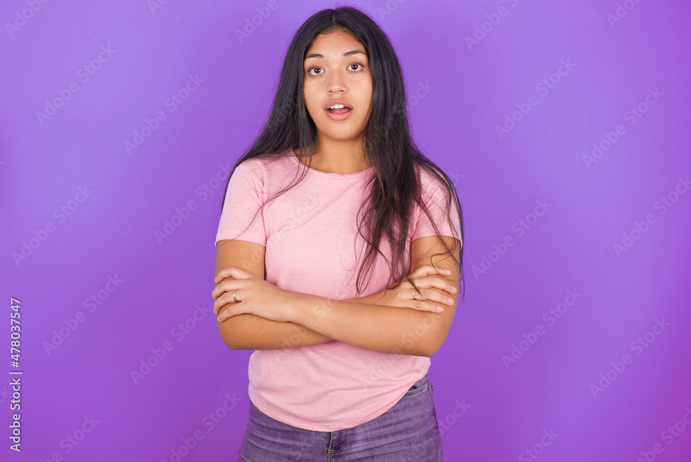 Shocked embarrassed Hispanic brunette girl wearing pink t-shirt over purple background keeps mouth widely opened. Hears unbelievable novelty stares in stupor