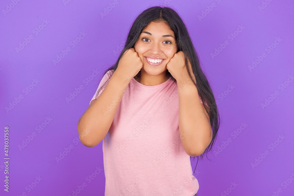 Happy Hispanic brunette girl wearing pink t-shirt over purple background keeps fists on cheeks smiles broadly and has positive expression being in good mood Happy Hispanic brunette girl wearing pink t-shirt over purple background keeps fists on cheeks smiles broadly and has positive expression being in good mood