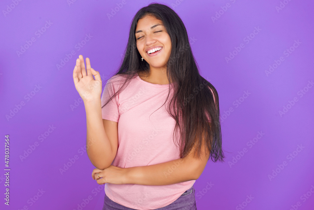 Fototapeta premium Overjoyed successful Hispanic brunette girl wearing pink t-shirt over purple background raises palm and closes eyes in joy being entertained by friends