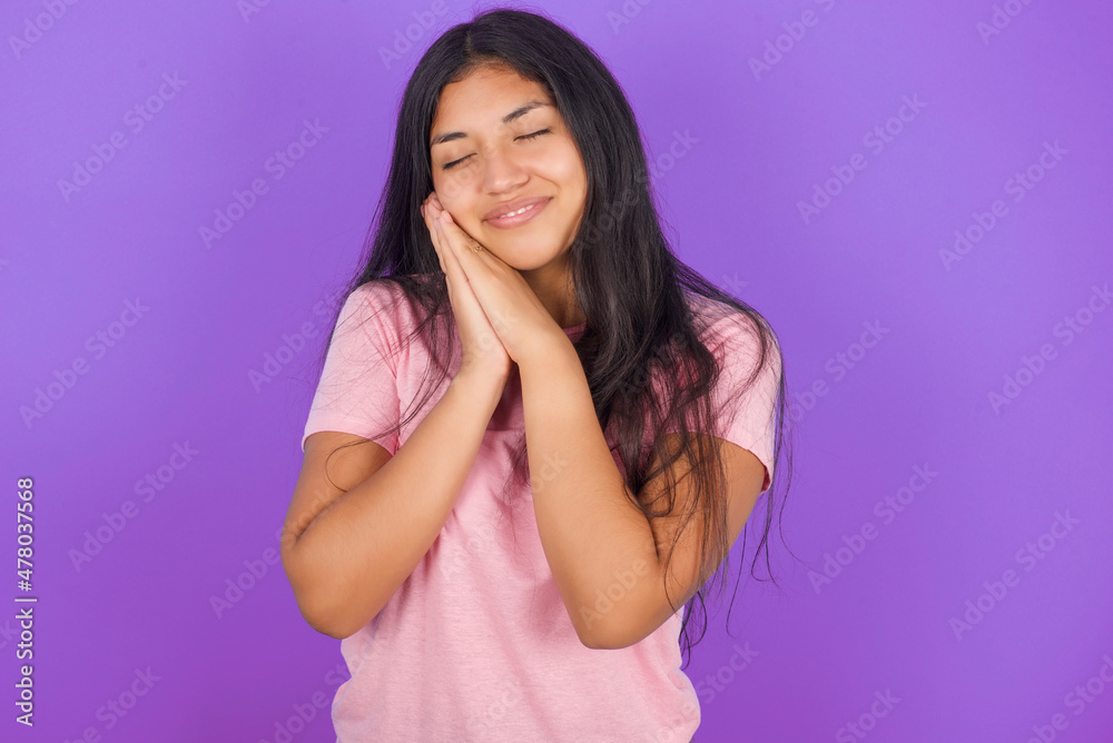 Hispanic brunette girl wearing pink t-shirt over purple background leans on pressed palms closes eyes and has pleasant smile dreams about something Hispanic brunette girl wearing pink t-shirt over purple background leans on pressed palms closes eyes and has pleasant smile dreams about something