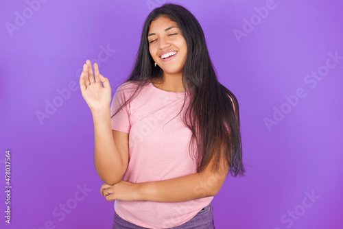 Overjoyed successful Hispanic brunette girl wearing pink t-shirt over purple background raises palm and closes eyes in joy being entertained by friends