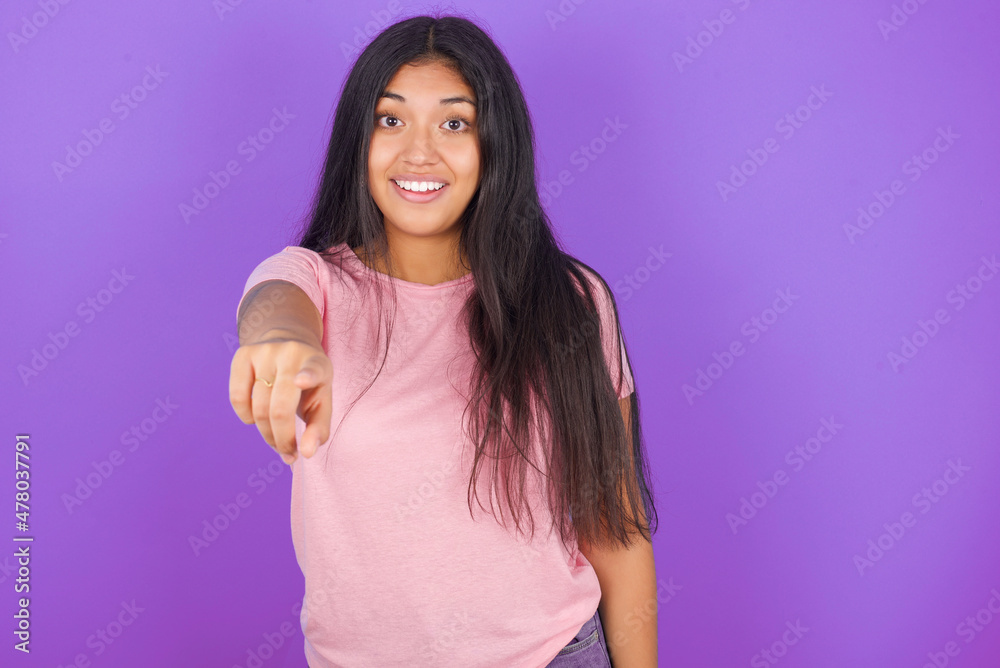 Excited positive Hispanic brunette girl wearing pink t-shirt over purple background points index finger directly at you, sees something very funny. Wow, amazing