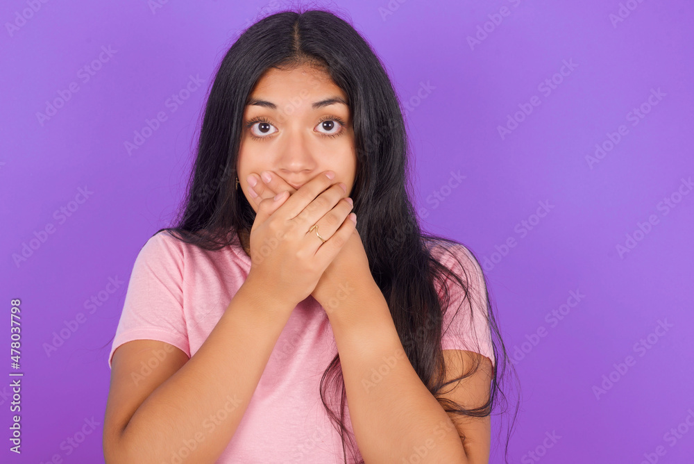 Stunned Hispanic brunette girl wearing pink t-shirt over purple background covers both hands on mouth, afraids of something astonishing Stunned Hispanic brunette girl wearing pink t-shirt over purple background covers both hands on mouth, afraids of something astonishing