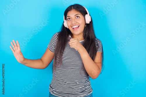 Happy hispanic girl wearing striped t-shirt standing over blue background sings favourite song keeps hand near mouth as if microphone wears wireless headphones, listens music