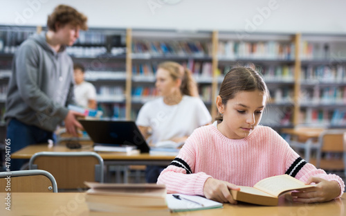 Young girl sitting at table in reading room in library and doing homework.