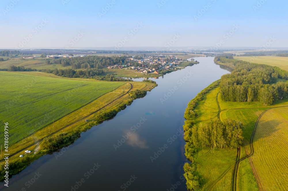 Aerial view of a wide river and a village in the distance among fields in Siberia in summer