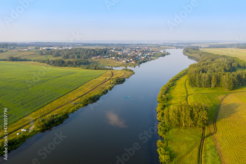 Aerial view of a wide river and a village in the distance among fields in Siberia in summer