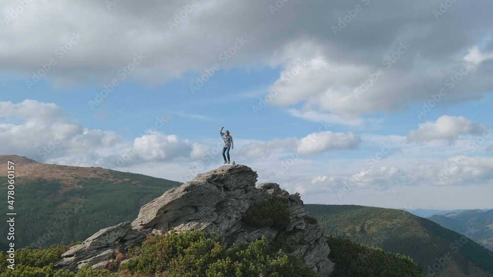 Happy man jumps on top of mountain rock on sunny day. Background of blue sky, clouds. Young man alone has conquered top and jumps on cliff with happiness. Success achievement concept