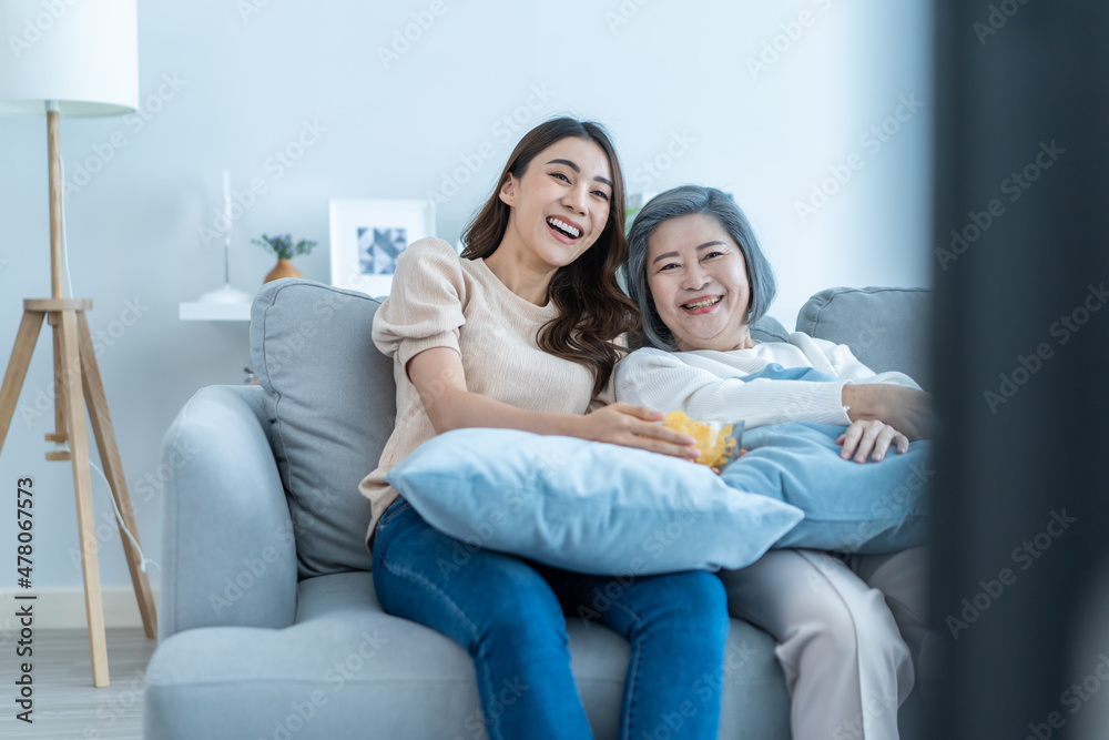 Asian senior mature woman and daughter sit on sofa watch funny movie.