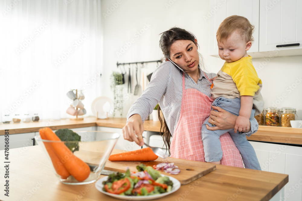 Caucasian busy mother doing housework with baby boy toddler in kitchen