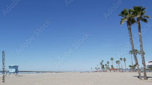 Tropical palm trees on white sandy beach by sea water waves, pacific ocean coast, Oceanside California USA. Blue sky and lifeguard tower. Life guard watchtower hut, summertime shore. Los Angeles vibes