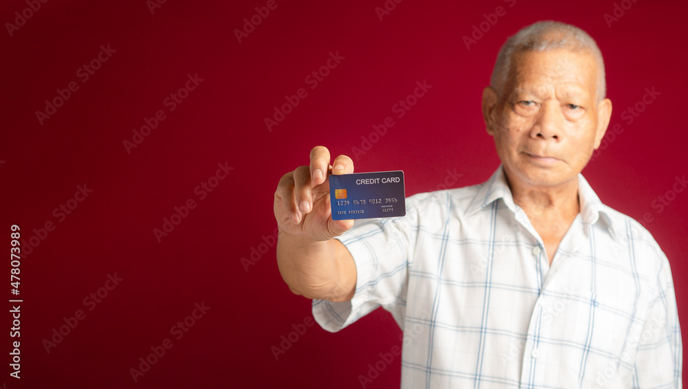 Senior man holding a blue credit card and looking at the camera with a smile while standing over a red background. Selective focus Senior man holding a blue credit card and looking at the camera with a smile while standing over a red background. Selective focus