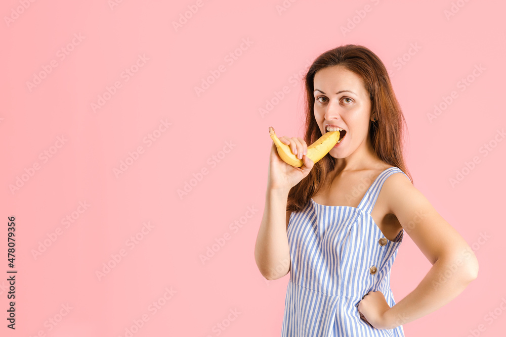 A brunette woman bites a yellow banana with a peel standing on a pink background
