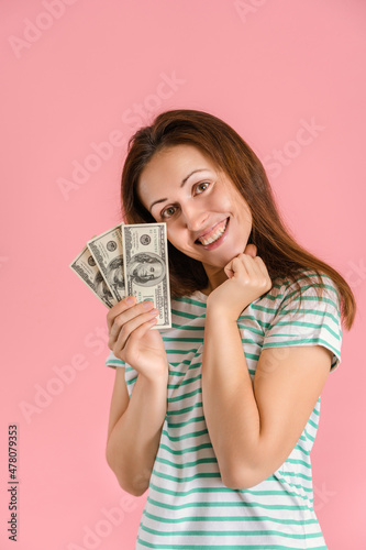 A joyful middle-aged girl smiles holding a fan of hundred-dollar bills in her hands standing on a pink background