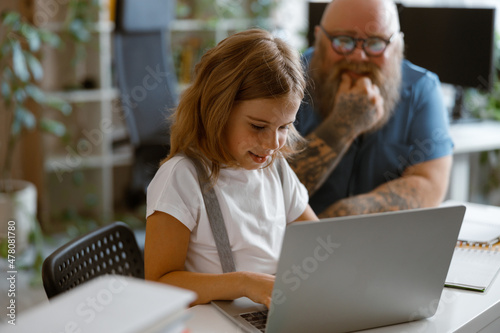 Smiling little girl types on laptop while father sits nearby at table in light room