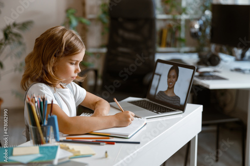 Little girl studies distantly at videocall via laptop at table in light room