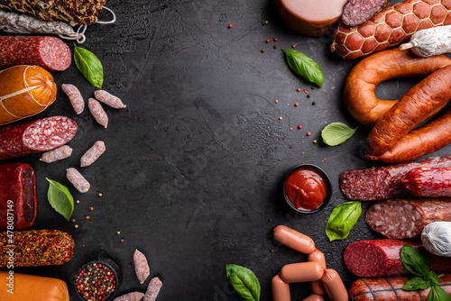 Set of different types of sausages, salami and smoked meat with basil and spices on a black stone background. Top view.