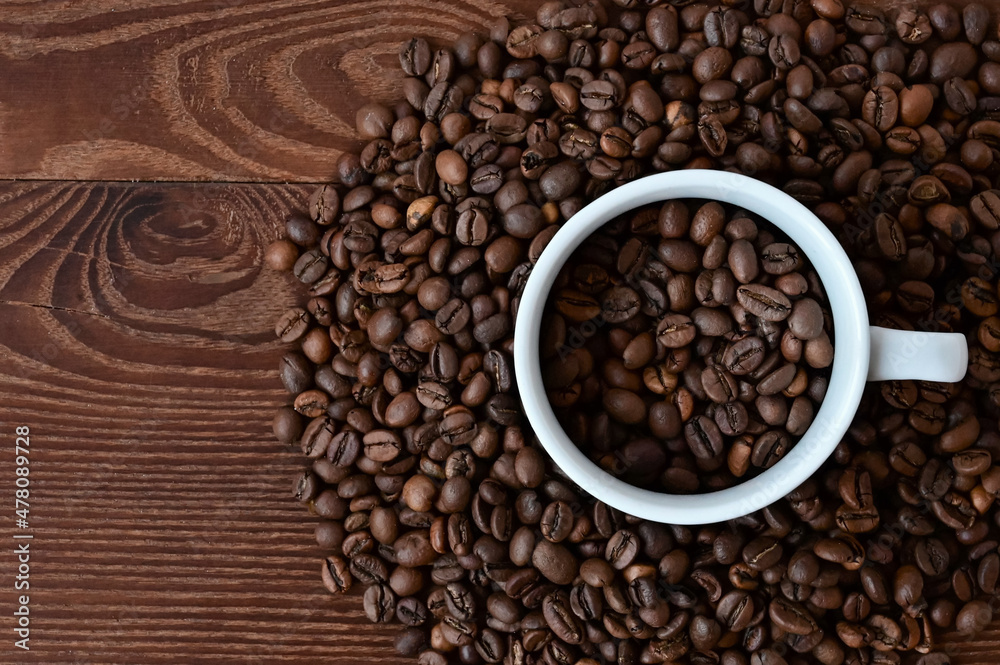 white cup filled with coffee beans on coffee background. Coffee beans on wooden table. Flat layout with top view. Copy space