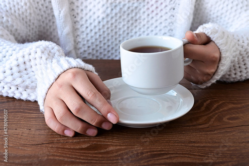 Close-up of girl's hands in a warm knitted white sweater holding cup of tea on wooden table. Winter holidays and lifestyle concept