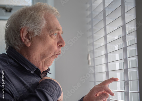 Obraz na plátně Senior man with funny facial expression spying through window blinds