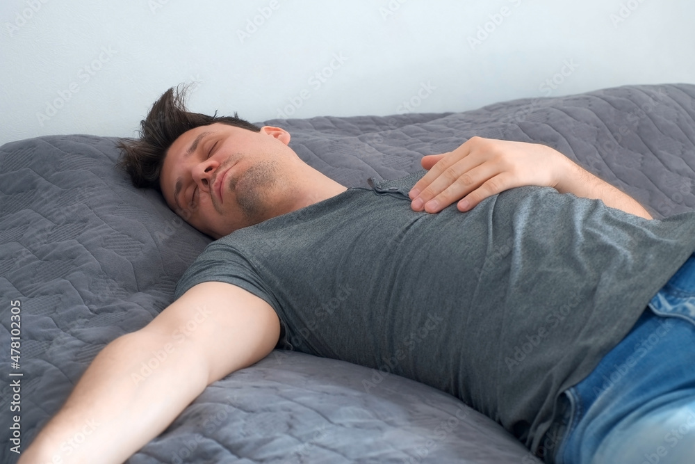 Young man is sleeping in bed wearing in jeans and t-shirt after party. Man with morning hangover at home. He sleeps on the unmade bed. The life of a person with bad habits, alcoholism. Young man is sleeping in bed wearing in jeans and t-shirt after party. Man with morning hangover at home. He sleeps on the unmade bed. The life of a person with bad habits, alcoholism.