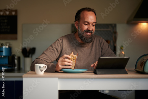 Positive bearded adult male sitting at table with cup of drink and bread toast browsing modern digital tablet in kitchen