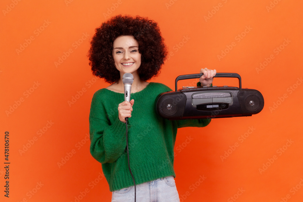 Smiling woman with Afro hairstyle wearing green casual style sweater standing holding record player and microphone in hands, looking at camera. Indoor studio shot isolated on orange background. Smiling woman with Afro hairstyle wearing green casual style sweater standing holding record player and microphone in hands, looking at camera. Indoor studio shot isolated on orange background.