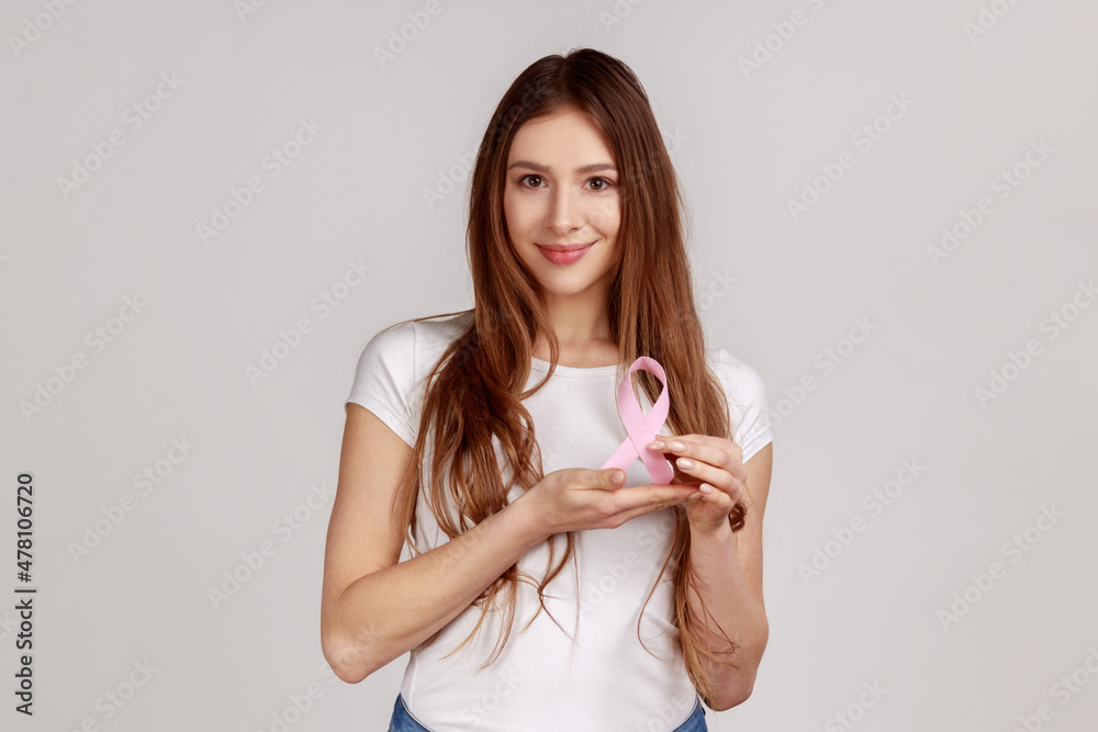 Women's health care, timely diagnosis. Happy smiling optimistic woman holding pink ribbon, symbol of breast cancer awareness, wearing white T-shirt. Indoor studio shot isolated on gray background. Women's health care, timely diagnosis. Happy smiling optimistic woman holding pink ribbon, symbol of breast cancer awareness, wearing white T-shirt. Indoor studio shot isolated on gray background.