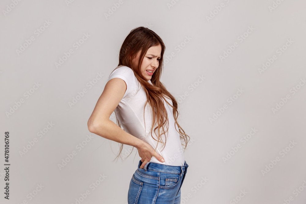 Portrait of young adult unhealthy woman suffering from lower back pain, pinched nerve or kidney inflammation, wearing white T-shirt. Indoor studio shot isolated on gray background. Portrait of young adult unhealthy woman suffering from lower back pain, pinched nerve or kidney inflammation, wearing white T-shirt. Indoor studio shot isolated on gray background.