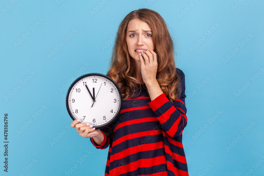 Confused woman wearing striped casual style sweater, holding wall clock in hands, biting nails, looking at camera with nervous expression, deadline. Indoor studio shot isolated on blue background. Confused woman wearing striped casual style sweater, holding wall clock in hands, biting nails, looking at camera with nervous expression, deadline. Indoor studio shot isolated on blue background.