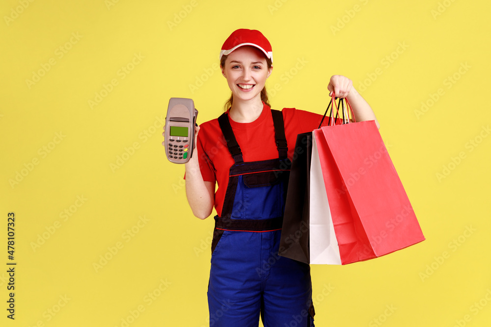 Portrait of happy delivery woman standing with shopping bags in hands and terminal for contactless payment, looking at camera, wearing overalls. Indoor studio shot isolated on yellow background.