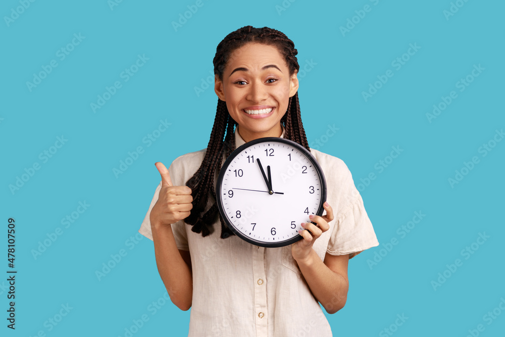 Positive happy woman with dreadlocks showing thumb up holding big wall clock, looking at camera with toothy smile, time management, wearing white shirt. Indoor studio shot isolated on blue background. Positive happy woman with dreadlocks showing thumb up holding big wall clock, looking at camera with toothy smile, time management, wearing white shirt. Indoor studio shot isolated on blue background.