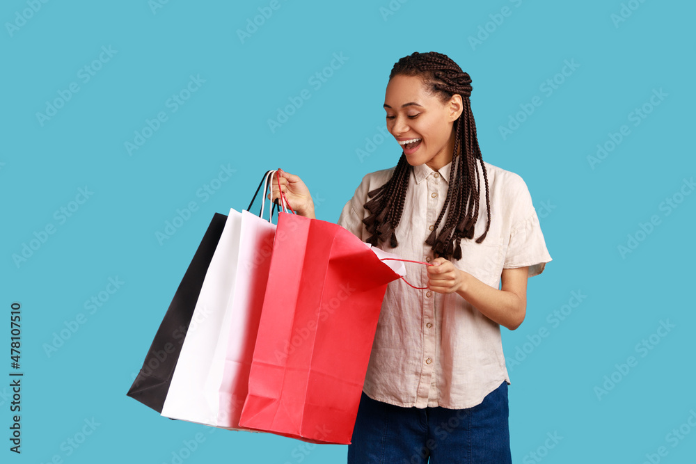 Excited smiling woman with black dreadlocks looking inside shopping bags, having positive expression, being satisfied with purchases.Indoor studio shot isolated on blue background. Excited smiling woman with black dreadlocks looking inside shopping bags, having positive expression, being satisfied with purchases.Indoor studio shot isolated on blue background.