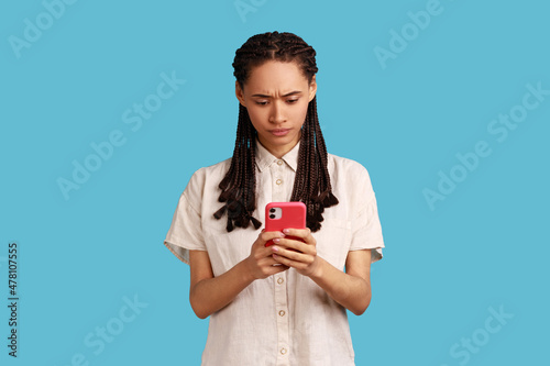 Serious looking woman with dreadlocks reads message on modern mobile phone, surfes social media, has concentrated look in display, updates information. Indoor studio shot isolated on blue background.