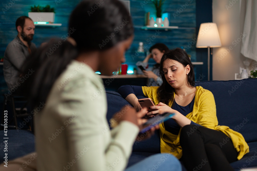 Multi-ethnic friends sitting on couch holding their modern smartphone surfing on internet, reading news during relaxing party. Group of people enjoying spending time together. Friendship concept