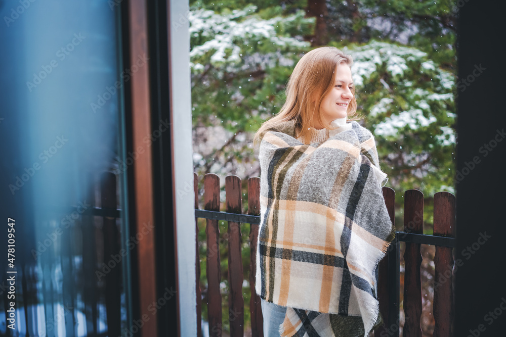 Young happy girl blonde woman on the balcony of her house enjoying the snow and forest view wrapping herself in a checkered plaid Young happy girl blonde woman on the balcony of her house enjoying the snow and forest view wrapping herself in a checkered plaid
