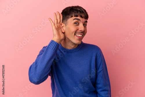 Young mixed race man isolated on pink background trying to listening a gossip.