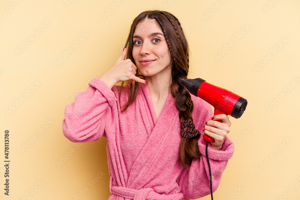 Young woman holding a hairdryer isolated on yellow background showing a mobile phone call gesture with fingers. Young woman holding a hairdryer isolated on yellow background showing a mobile phone call gesture with fingers.