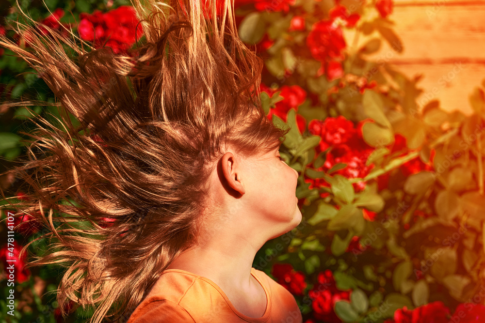 Naklejka premium A child's long hair grows in strong winds. Girl's hairstyle close-up on a background of roses in the garden. A child plays and indulges outdoor on a summer day.