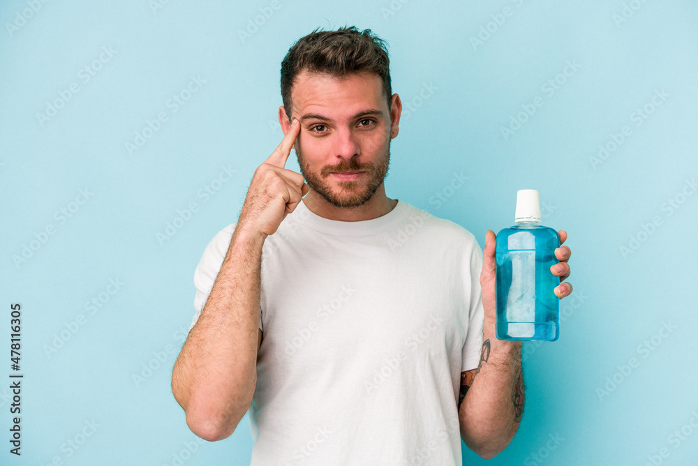 Young caucasian man holding mouthwash isolated on blue background pointing temple with finger, thinking, focused on a task. Young caucasian man holding mouthwash isolated on blue background pointing temple with finger, thinking, focused on a task.
