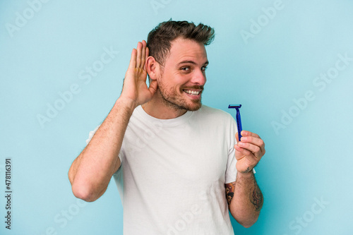 Young caucasian man shaving his beard isolated on blue background trying to listening a gossip.