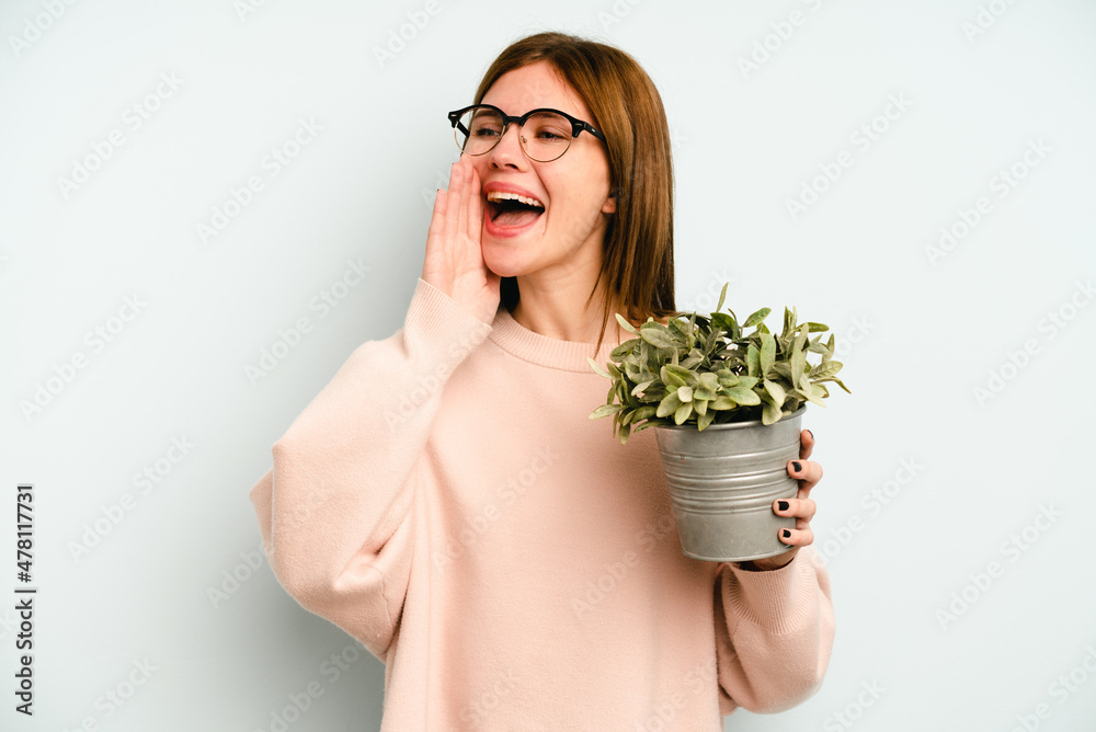 Young English woman holding a plant isolated on blue background shouting and holding palm near opened mouth. Young English woman holding a plant isolated on blue background shouting and holding palm near opened mouth.