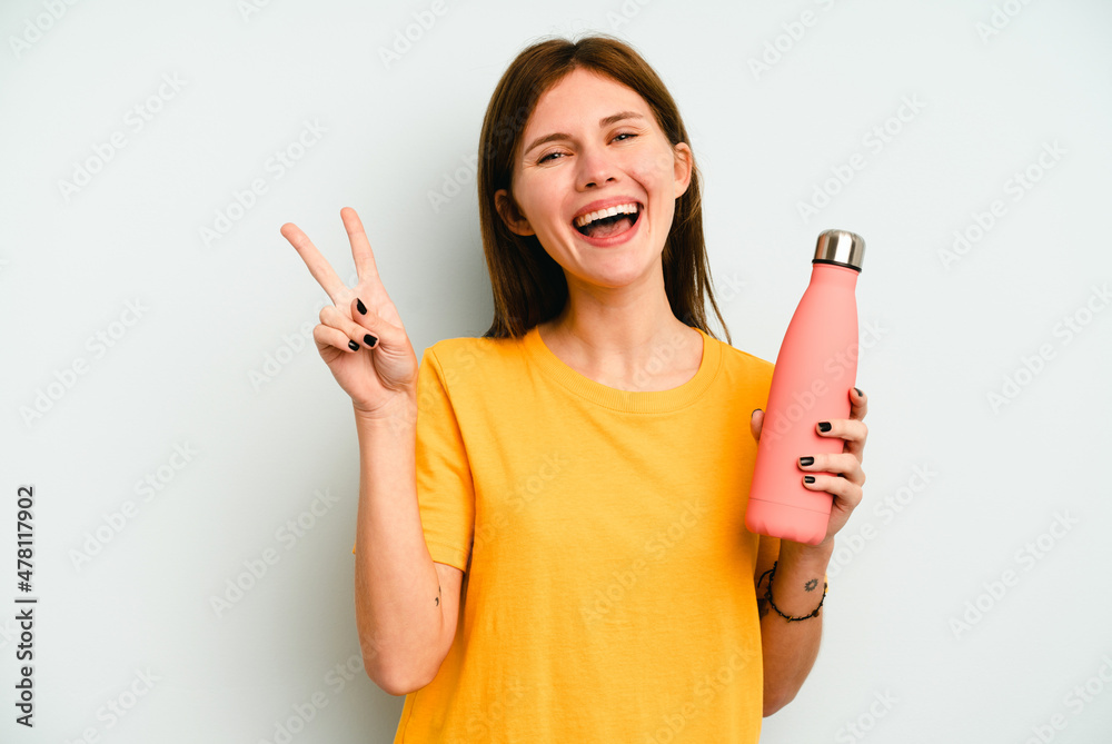 Young English woman holding a thermo isolated on blue background joyful and carefree showing a peace symbol with fingers. Young English woman holding a thermo isolated on blue background joyful and carefree showing a peace symbol with fingers.