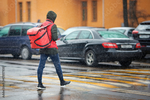 Food delivery courier with red backpack delivers food in winter. Courier works during heavy snowfall, deliver food orders to customers. Deliveries work in difficult working conditions in any weather