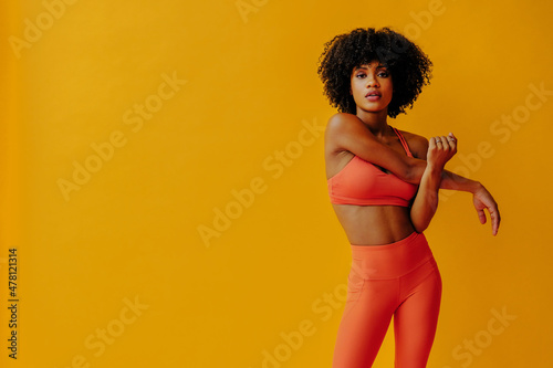 attractive young adult fit African American woman in sportswear stretching isolated on orange background