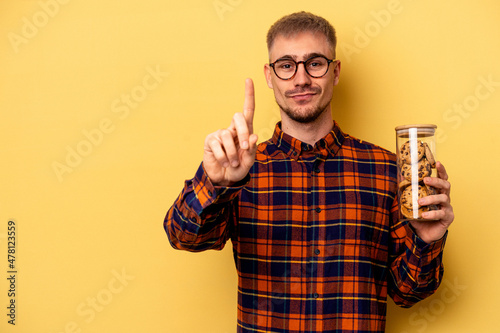 Young caucasian man holding cookies jar isolated on yellow background showing number one with finger.